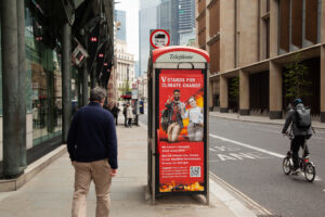 A red phone booth is positioned on a sidewalk in downtown London's financial district. A man dressed in khakis and a navy sweater walks by, his back facing the camera. On the street next to the phone booth, a cyclist is riding by. On the phone booth is a poster calling out Vanguard which reads "V stands for climate change. We haven't changed much since 1975. But the climate has, thanks to our steadfast investments in coal, oil, and gas." There are two happy looking people pictured on the poster each making V shapes with their first two fingers, mimicking a Vanguard marketing campaign.