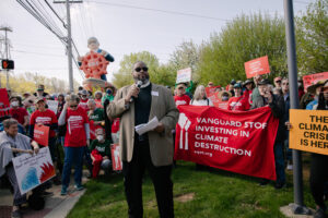 A photo of Bishop Dwayne Royster from Power speaking into a microphone in front of a gathering of 50+ protestors. In the background is an inflatable of Vanguard CEO Tim Buckley in a ship that's wreathed in painted flames, clutching a red steering wheel with S.O.S. on the front. At the front of the crowd, two elderly protestors hold a red banner that reads “Vanguard stop investing in climate destruction” with an illustration of two smokestacks on the left. Other protestors hold signs reading “don’t torch earth for your retirement” and “the climate crisis is here”
