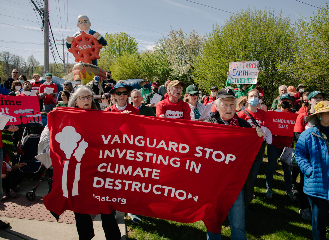 A gathering of 50+ protestors. In the background is an inflatable of Vanguard CEO Tim Buckley in a ship that's wreathed in painted flames, clutching a red steering wheel with S.O.S. on the front. At the front, two elderly protestors hold a red banner that reads “Vanguard stop investing in climate destruction” with an illustration of two smokestacks on the left. Other protestors hold signs reading "It's time to cut carbon" and "Vanguard S.O.S.” and “Don’t torch Earth for your retirement.”