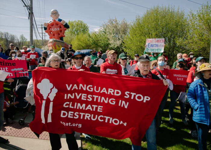 A gathering of 50+ protestors. In the background is an inflatable of Vanguard CEO Tim Buckley in a ship that's wreathed in painted flames, clutching a red steering wheel with S.O.S. on the front. At the front, two elderly protestors hold a red banner that reads “Vanguard stop investing in climate destruction” with an illustration of two smokestacks on the left. Other protestors hold signs reading "It's time to cut carbon" and "Vanguard S.O.S.” and “Don’t torch Earth for your retirement.”