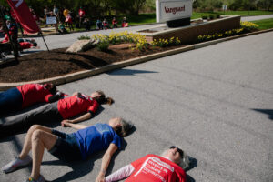 Four protestors lie in the road, blocking the driveway of Vanguard headquarters. In the background you can see the Vanguard sign in the median surrounded by yellow flowers. Three of the four protestors wear red shirts reading “Vanguard invests in climate destruction.” In the background more protestors are visible, holding signs.