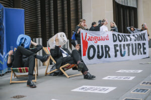Two demonstrators dressed in suits sit on lawn chairs looking relaxed, holding champagne flutes, wearing plastic oil barrels on their heads with silly-looking faces painted on them. One of them is holding his middle finger up to the camera. They sit on a public street in London next to other demonstrators holding a large sign that reads "Vanguard: Don't Sink Our Future". Additional protest signs are plastered to the ground.