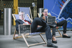 Two demonstrators lounge in deck chairs holding chalkboards that read "Tim Buckley CEO + Chair Vanguard" and "Sean Hegarty MD Europe." The two demonstrators also wear facemasks of each Tim Buckley and Sean Hegarty. The person representing Hegarty holds a cigar. In the background, part of a painting depicting the Titanic on a huge cloth banner is visible