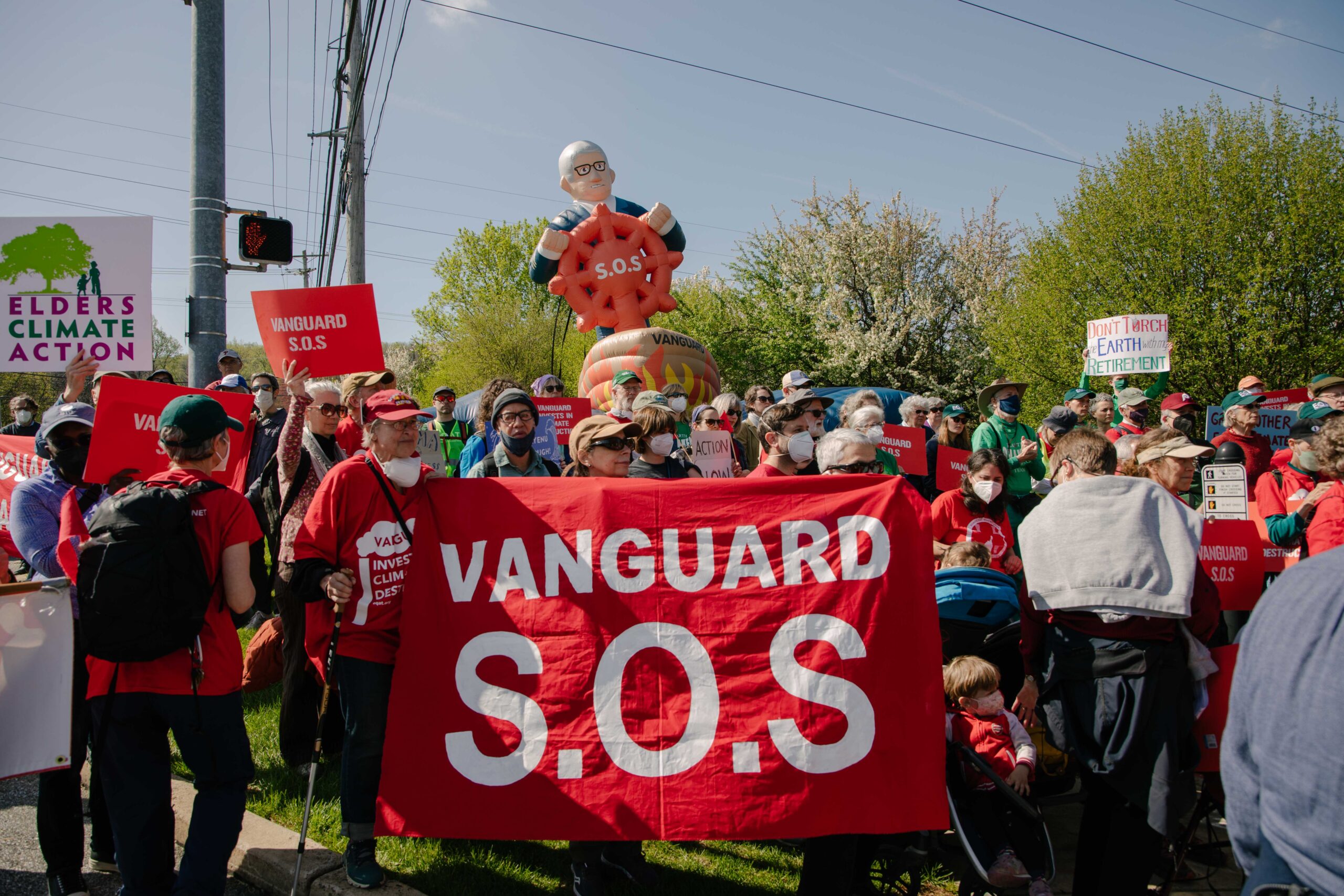 A group of 50+ protestors gather carrying signs. In the front of the crowd protestors hold a red banner with white text reading "Vanguard S.O.S." In the background is an inflatable of Vanguard CEO Tim Buckley in a ship that's wreathed in painted flames, clutching a red steering wheel with S.O.S. on the front.