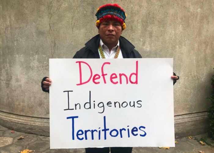 A man who is a member of the Achuar people holds a sign that says 'Defend Indigenous Territories'