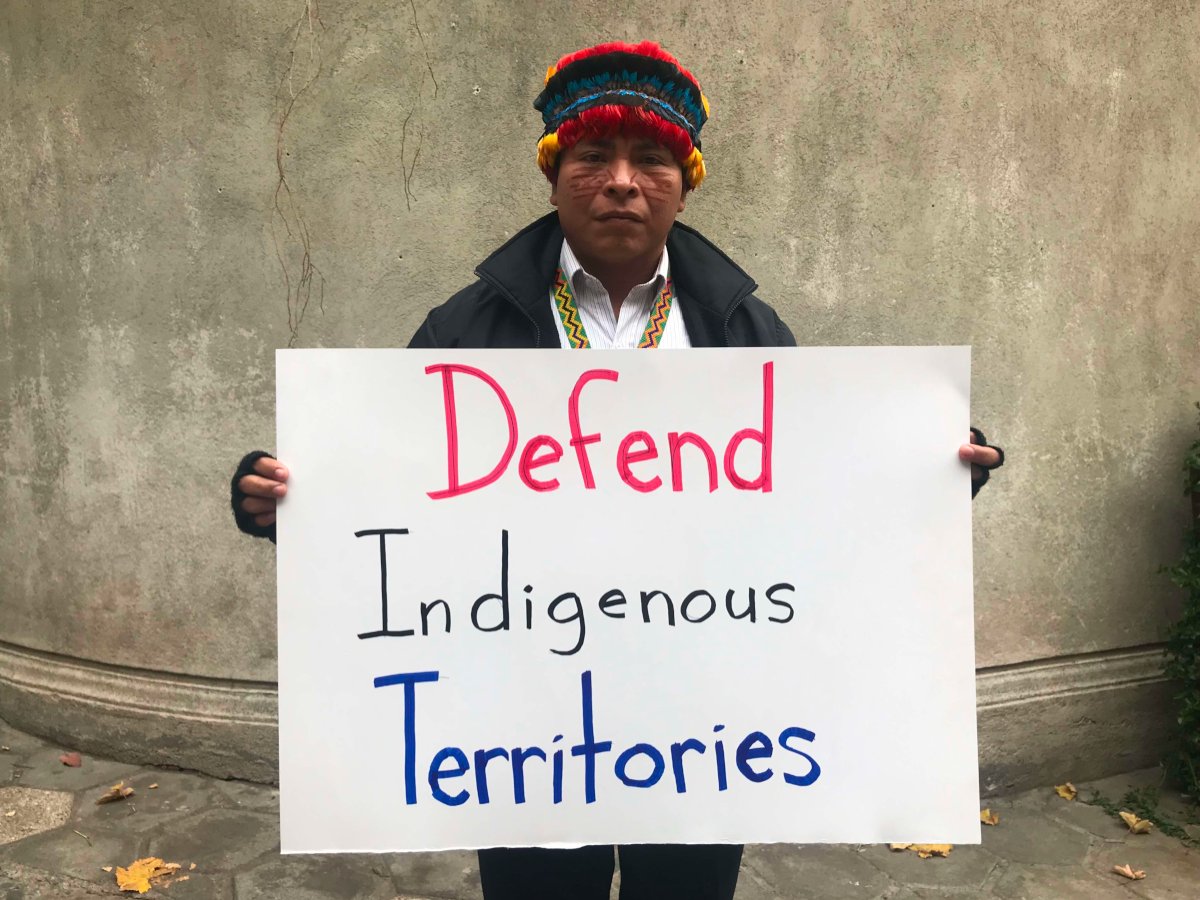 A man who is a member of the Achuar people holds a sign that says 'Defend Indigenous Territories'