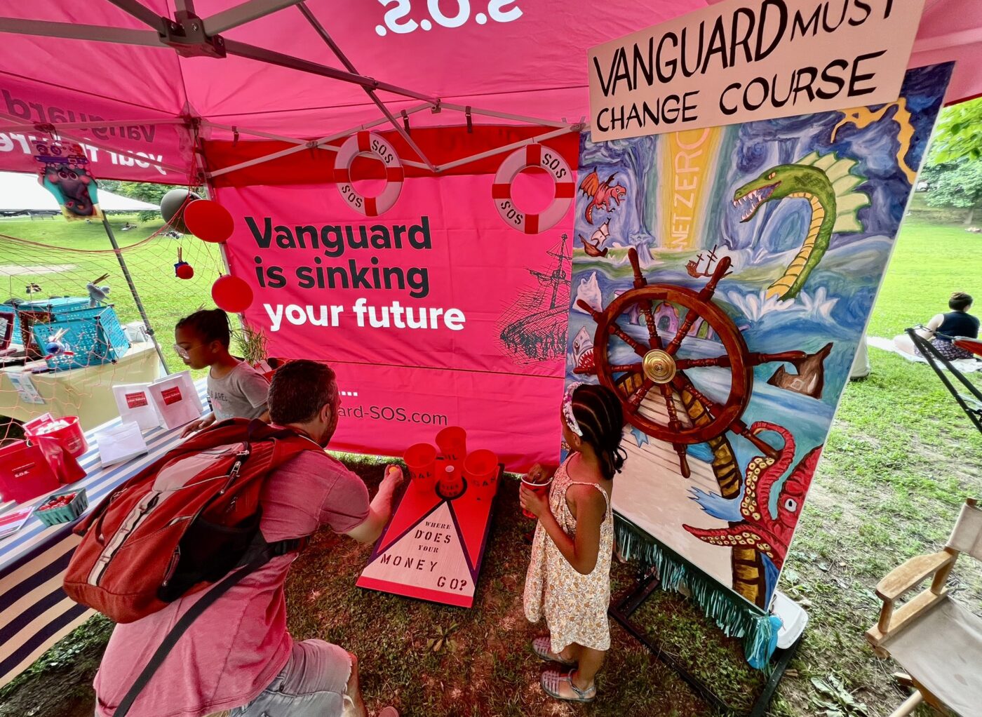 Community members play nautical themed carnival games that highlight Vanguard's climate problem at a community festival in Philadelipha. They are standing under a large banner that reads "Vanguard is sinking your future."