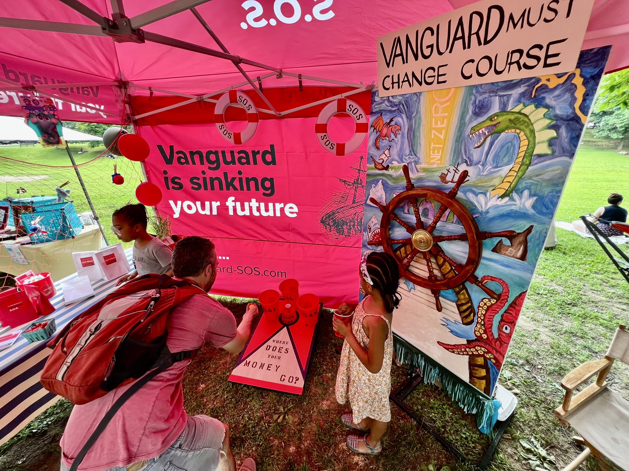 Community members play nautical themed carnival games that highlight Vanguard's climate problem at a community festival in Philadelipha. They are standing under a large banner that reads "Vanguard is sinking your future."
