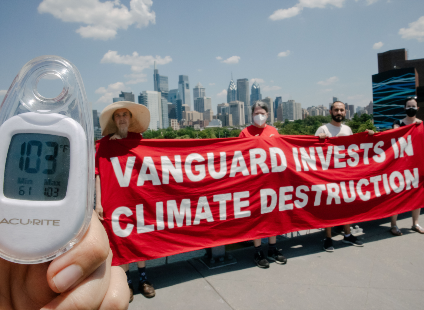 Activists hold a sign that reads "Vanguard invests in climate destruction" during a heat wave in Philadelphia. Someone is showing a thermometer that reads 103 degrees F.